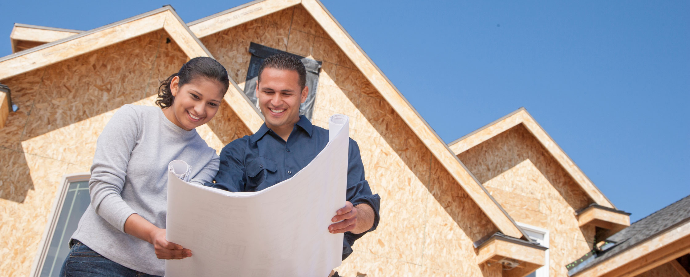 Photo of couple looking at a set of blueprints with a house under construction behind them